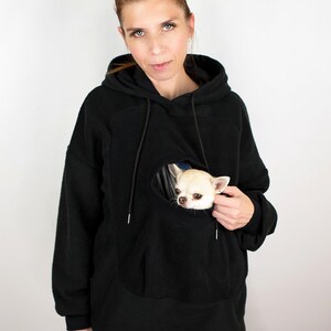 May include: A black fleece hoodie with a large pocket on the front. The pocket has a hole in it, and a small white dog is peeking out of the hole.