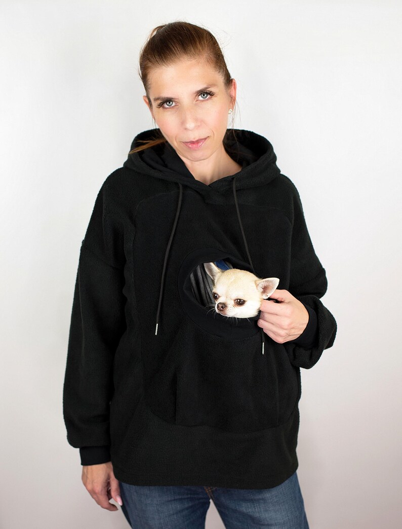 May include: A black fleece hoodie with a large pocket on the front. The pocket has a hole in it, and a small white dog is peeking out of the hole.