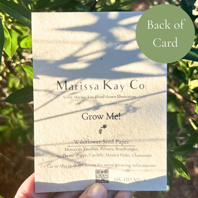 May include: A seed paper card with the text "Marissa Kay Co" and "Grow Me!". The card lists wildflower seeds: Moroccan Toadflax, Petunia, Beardtongue, Daisy, Thyme, Poppy, Catchfly, Maiden Pinks, and Chamomile. The card is held in front of a green leafy background.