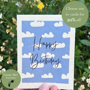 Könnte beinhalten: Handgemachte Geburtstagskarte mit blauem Himmel und Wolkenmuster. Die Worte "Happy Birthday" sind in eleganter Schrift geschrieben. Die Karte besteht aus Saatpapier und hat ein natürliches, strukturiertes Aussehen. Die Karte wird vor einem grünen Hintergrund gehalten.