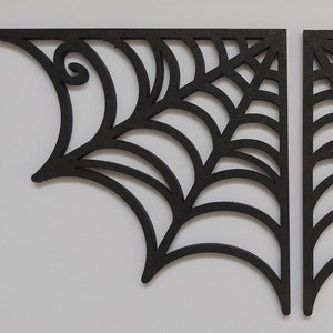 Könnte beinhalten: Zwei schwarze, dekorative Eckregalhalterungen mit Spinnennetz-Design. Jede Halterung hat eine gebogene, fächerartige Form mit einer Reihe von gewölbten Linien und einer dekorativen Verzierung oben. Die Halterungen sind aus dunklem Material.