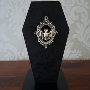 May include: A silver bat pendant with detailed wings, set in an oval frame, displayed on a black, coffin-shaped stand. The design has a gothic or Halloween theme. The background features a patterned wallpaper.