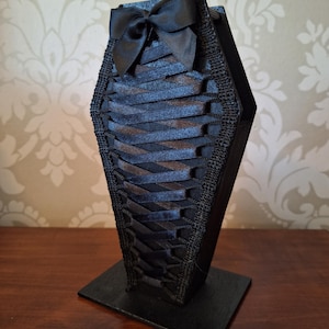 May include: A black coffin-shaped decorative box with a satin bow at the top. The box is decorated with woven black ribbon and stands on a square base. The design has a gothic or Victorian aesthetic.