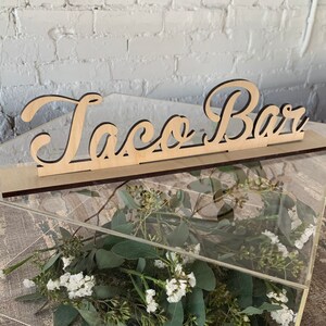 May include: A clear acrylic box with a wooden sign that reads "Taco Bar". The sign is made of light-colored wood with a dark border. The box contains greenery and white flowers, perfect for a party or event.