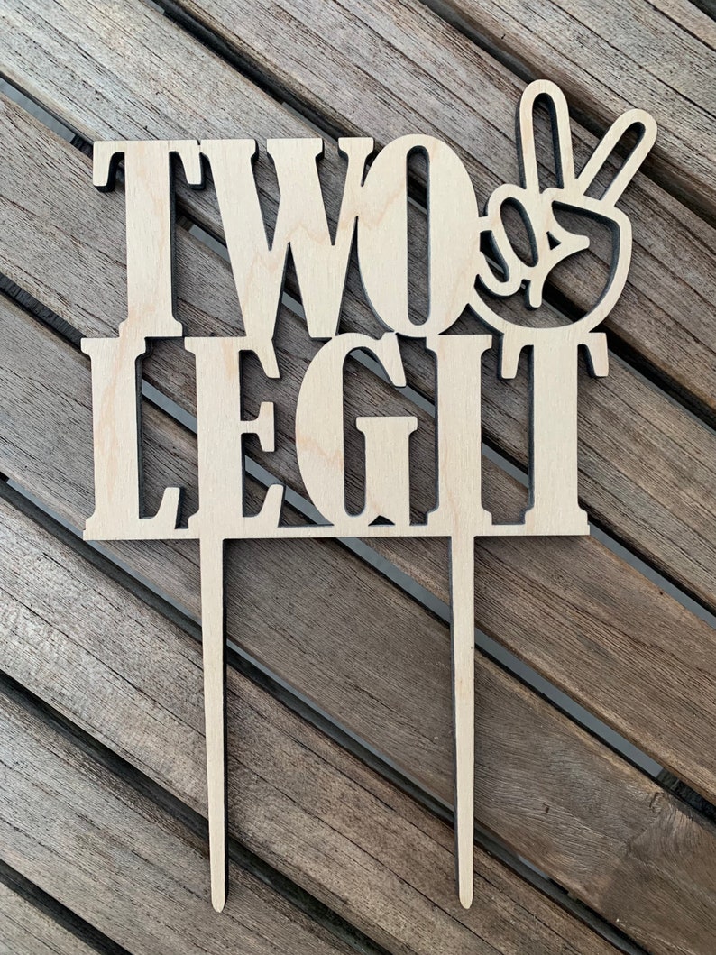 May include: Wooden cake topper with the words "TWO LEGIT" and a peace sign. The topper is designed for a birthday cake.