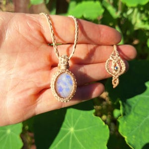 May include: A macrame necklace featuring an oval moonstone pendant with a blue iridescent sheen, suspended from a braided beige cord, accompanied by a matching small macrame earring or charm with a central silver-toned metal bead.