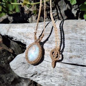 May include: A macrame pendant necklace featuring an oval-shaped iridescent moonstone cabochon encased in light tan woven cord. The necklace includes a matching tan cord with a decorative macrame knot detail and a small metallic bead accent.