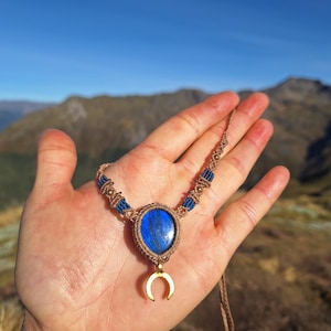 May include: A macrame necklace featuring a central teardrop-shaped blue labradorite stone encased in tan cord. A gold-toned crescent moon charm hangs below the stone. The cord is accented with blue thread segments and small metallic beads.