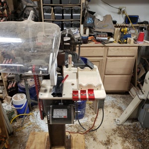 Op de afbeelding: Een close-up van een houtbewerkingsgereedschap met een doorzichtige kunststof beschermkap, een rode hendel en een grijze slijpschijf. Het gereedschap is gemonteerd op een houten basis met een zwarte motor en een rood elektrisch snoer. Er zijn drie rode klemmen op de basis.