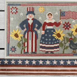 May include: A cross-stitch embroidery featuring a patriotic scene with a man and woman in red, white, and blue clothing, sunflowers, and a black rooster. The scene is framed with a red, white, and blue border.