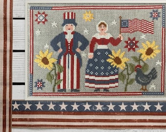 May include: A cross-stitch embroidery depicting a patriotic scene with a man and woman in red, white and blue clothing, sunflowers, and a black rooster. The scene is framed with a red, white and blue border.