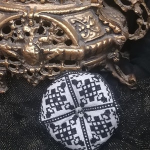 Puede incluir: Un alfiletero de tela con estampado en blanco y negro con un bot&oacute;n en el centro. El alfiletero est&aacute; sobre una superficie de tela negra.