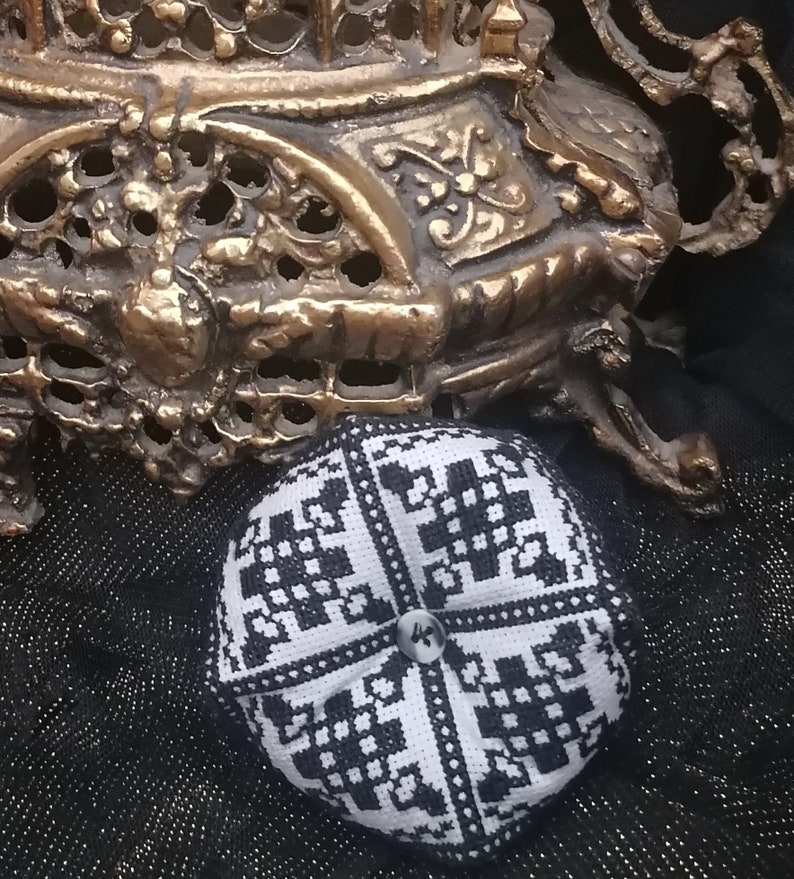 Puede incluir: Un alfiletero de tela con estampado en blanco y negro con un bot&oacute;n en el centro. El alfiletero est&aacute; sobre una superficie de tela negra.