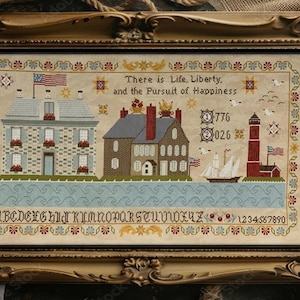Op de afbeelding: Een ingelijste kruissteek met een kustlandschap met gebouwen, een vuurtoren en een zeilboot. Het kunstwerk bevat de tekst "There is Life, Liberty, and the Pursuit of Happiness" en de nummers "776" en "026". De lijst is sierlijk en goudkleurig.