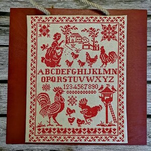 May include: A cross-stitch sampler with a red and white color scheme. The sampler features a rooster, a hen, chicks, a birdhouse, a house, trees, a woman, and the alphabet and numbers.