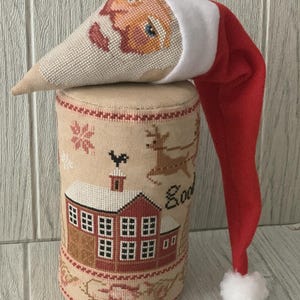 May include: A festive Christmas decoration. It features a red Santa hat with a white pom-pom and a cross-stitched Santa face. The cylindrical base is beige and decorated with cross-stitched images of a house, reindeer, and snowflakes.