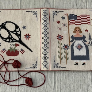 Puede incluir: Una aguja de punto de tela blanca con una bandera estadounidense roja, blanca y azul, una mujer con un vestido azul y una silueta negra de una jaula para pájaros. La aguja de punto está enmarcada con un borde negro y tiene un cordón rojo con dos borlas rojas.