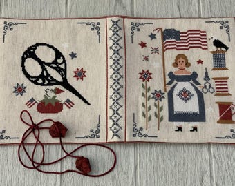 May include: Cross-stitch textile with patriotic themes. Features a large black pair of scissors, a woman holding an American flag, and various sewing-related elements. Includes red, white, and blue accents, and a decorative border. A red cord with two decorative pieces is in the foreground.