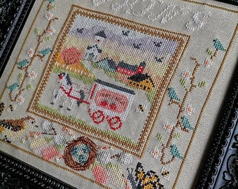 May include: A cross-stitch embroidery framed in a black ornate frame. The design features a country scene with a red barn, a horse-drawn wagon, and a bird's nest with eggs. The text "APRIL" is stitched in white at the top of the design.