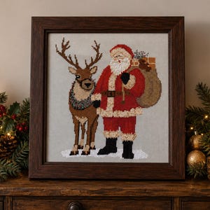 Può includere: Un ricamo a punto croce incorniciato raffigurante Babbo Natale in abito rosso con barba bianca in piedi accanto a una renna marrone. Babbo Natale porta un grande sacco beige sulla spalla. Il disegno è su uno sfondo in tessuto grigio chiaro.