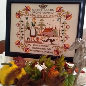 May include: A framed cross-stitch embroidery with a woman in a red dress standing in front of a house. The embroidery includes the text "Be on your guard stand firm in the faith be courageous, be strong".