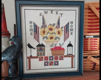 May include: A framed cross-stitch embroidery featuring a scene of a table with a vase of sunflowers, a bowl of apples, a small house, and a lantern. The text "SWEET HOME" is stitched above the scene.