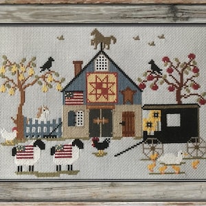 May include: A cross-stitch embroidery depicting a quaint farmhouse with a red, white, and blue quilt hanging on the side. The farmhouse is surrounded by trees, a black horse-drawn carriage, and various farm animals, including chickens, sheep, and ducks. The scene is framed in a rustic wooden frame.