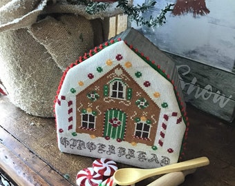 Op de afbeelding: Een kruissteek van een peperkoekhuis met de woorden "Gingerbread" eronder gestikt. Het huis is versierd met snoepstokken, dropjes en een groene deur. Er liggen twee pepermuntjes, een houten lepel en een deegroller op een houten tafel.