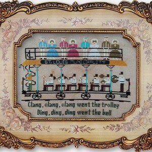 Puede incluir: Un bordado de punto de cruz enmarcado que representa un tranvía vintage con pasajeros. El tranvía está etiquetado como "ST. LOUIS MUNI" y el pie de foto dice "Clang, clang, clang went the trolley Ding, ding, ding went the bell".