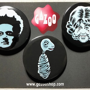 Op de afbeelding: Drie zwart-witte knoopsluitingen met verschillende afbeeldingen. De eerste knoopsluiting heeft een zwart-witte afbeelding van een persoon met een grote afro. De tweede knoopsluiting heeft een zwart-witte afbeelding van een wezen met een lange nek en een gewikkeld lichaam. De derde knoopsluiting heeft een zwart-witte afbeelding van een persoon met een groot, krullend kapsel. De tekst "GAZOO" is in wit op een rode achtergrond gedrukt.