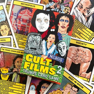 May include: A yellow, red and black box with the text "Cult Films 2 Trumps Card Game" on the front. The box is open and shows a variety of playing cards with images of characters from horror films. The cards have a yellow border and black text. The cards include characters from films like "The Texas Chainsaw Massacre" and "Society."