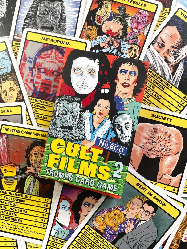 May include: A yellow, red and black box with the text "Cult Films 2 Trumps Card Game" on the front. The box is open and shows a variety of playing cards with images of characters from horror films. The cards have a yellow border and black text. The cards include characters from films like "The Texas Chainsaw Massacre" and "Society."