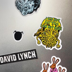K&ouml;nnte beinhalten: Eine Sammlung von Aufklebern mit verschiedenen Designs. Enth&auml;lt eine schwarz-wei&szlig;e Silhouette, einen Aufkleber mit dem Text "DAVID LYNCH" und mehrere Illustrationen im Cartoon-Stil. Ein Aufkleber zeigt eine Kreatur mit dem Text "I DID NOT SAY THIS I AM NOT HERE".