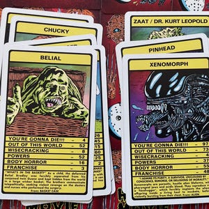 May include: A set of colourful trading cards featuring illustrations of horror movie monsters. Each card has a name, a statistic for "You're Gonna Die!!!", and a description of the monster's origin. The cards include characters like Chucky, Belial, Xenomorph, and Pinhead.