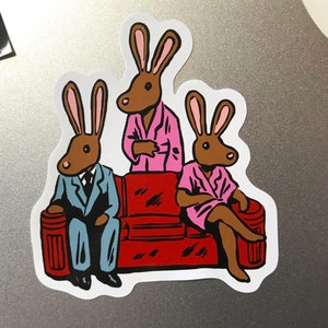 K&ouml;nnte beinhalten: Ein Aufkleber mit drei anthropomorphen Kaninchen. Eines tr&auml;gt einen blauen Anzug, ein anderes einen rosa Bademantel und das dritte einen rosa Bademantel. Sie sitzen auf einem roten Sofa. Die Kaninchen haben braune K&ouml;rper und rosa Ohren.