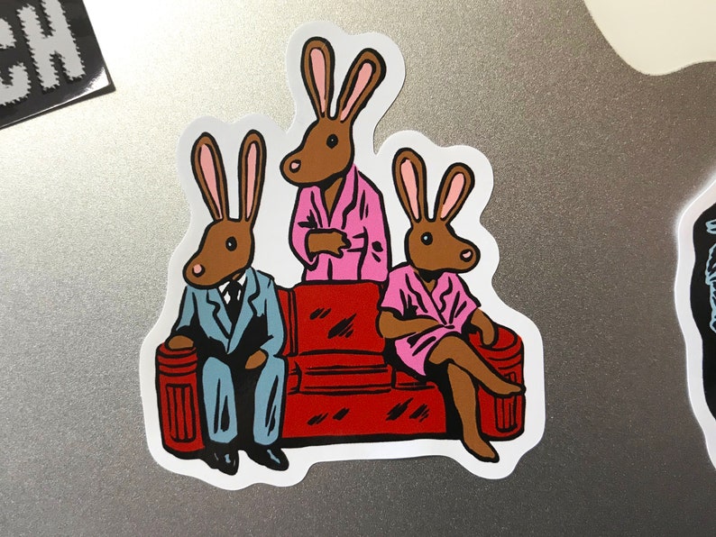 K&ouml;nnte beinhalten: Ein Aufkleber mit drei anthropomorphen Kaninchen. Eines tr&auml;gt einen blauen Anzug, ein anderes einen rosa Bademantel und das dritte einen rosa Bademantel. Sie sitzen auf einem roten Sofa. Die Kaninchen haben braune K&ouml;rper und rosa Ohren.