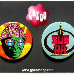 May include: Two round buttons with colorful graphics. One features a green face with orange glasses and a pink and yellow hat. The other has the text "SUN RA" in red and brown on a light blue background. A pink "GAZOO" logo is above.