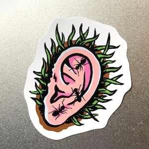 K&ouml;nnte beinhalten: Ein Aufkleber mit einem stilisierten Ohr mit Ameisen, die darin krabbeln. Das Ohr ist rosa mit schwarzen Umrissen, umgeben von gr&uuml;nen grasartigen Elementen. Die Ameisen sind schwarz. Der Aufkleber hat einen wei&szlig;en Rand und liegt auf einer grauen Oberfl&auml;che.