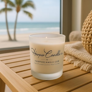 Może przedstawiać: Zapalone, matowe szklane świece z napisem "Harper Candle co." i "HARPERCANDLE.COM Hand Poured Coconut Wax Candles." Świeca stoi na drewnianej powierzchni z widoczną w tle sceną plażową.