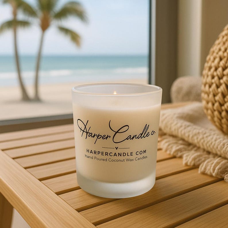 Może przedstawiać: Zapalone, matowe szklane świece z napisem "Harper Candle co." i "HARPERCANDLE.COM Hand Poured Coconut Wax Candles." Świeca stoi na drewnianej powierzchni z widoczną w tle sceną plażową.