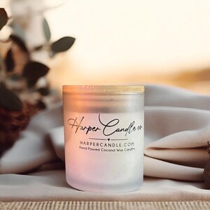 Może przedstawiać: Szklany słoik na świecę z matowego szkła z drewnianą pokrywką. Słoik ma etykietę z napisem "Harper Candle co." i "HARPERCANDLE.COM Hand-Poured Coconut Wax Candles." Świeca ma r&oacute;żowo-biały gradient. Tło jest rozmyte.