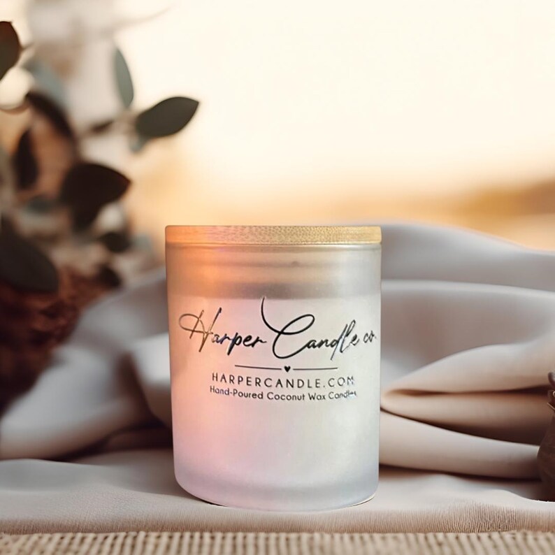 Może przedstawiać: Szklany słoik na świecę z matowego szkła z drewnianą pokrywką. Słoik ma etykietę z napisem "Harper Candle co." i "HARPERCANDLE.COM Hand-Poured Coconut Wax Candles." Świeca ma r&oacute;żowo-biały gradient. Tło jest rozmyte.