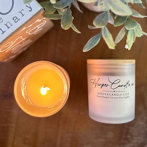 Może przedstawiać: Dwie zapalone świece, jedna w przezroczystym szkle, druga w matowym słoiku z drewnianą pokrywką. Słoik jest oznaczony "Harper Candle co." i "Hand-Poured Coconut Wax Candles". Świece stoją na ciemnej drewnianej powierzchni.
