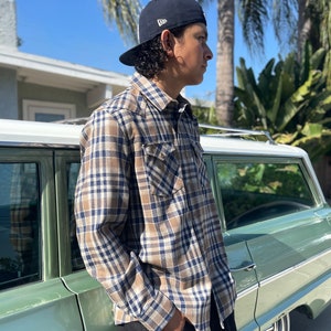 May include: A person wearing a blue and brown plaid shirt, black pants, and a black baseball cap stands next to a green vintage car.