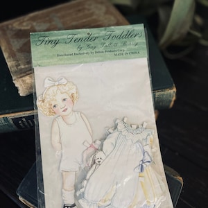 May include: A paper doll set featuring a blonde-haired girl wearing a white dress and holding a small white dog. The set includes a blue dress with a yellow sash and a white collar. The packaging includes the text "Tiny Tender Toddlers by Gay Talbott Boassy Distributed Exclusively by Delton Products Corp. MADE IN CHINA."