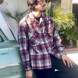 May include: A long-sleeved plaid shirt in shades of red, blue, and tan. The shirt has two chest pockets and a button-down front. The person is wearing black pants and a black undershirt. The shirt is leaning against a vintage green car.
