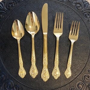 May include: A gold-colored flatware set, including a spoon, knife, and two forks, arranged on a black decorative plate. The utensils have ornate floral detailing on the handles. The plate has a raised, swirling pattern around the edge.