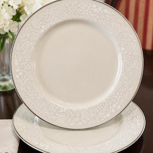 May include: Two round ceramic dinner plates featuring a smooth, light beige center and a wide rim decorated with a detailed white floral pattern. Each plate is finished with a thin, metallic silver edge along the outer circumference.