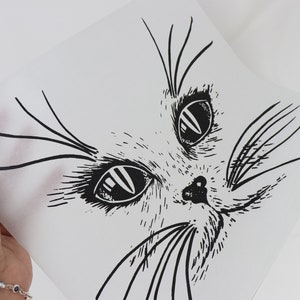 May include: Black and white line drawing of a cat's face on white paper. The cat's eyes are large and round, and its whiskers are long and thin.