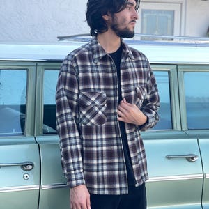 May include: A plaid flannel shirt in shades of brown, white, and blue. The shirt has a front pocket and a zipper closure. The person is wearing a black t-shirt underneath and black pants. The background includes a vintage green car.